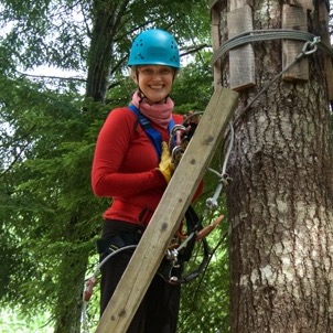 Nicole Lantz in protective climbing gear standing by a wooden and metal wire obstacle attached to a large tree