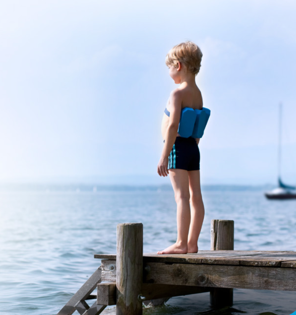 A young boy wearing a swim suit and float support, standing on a pier over the water