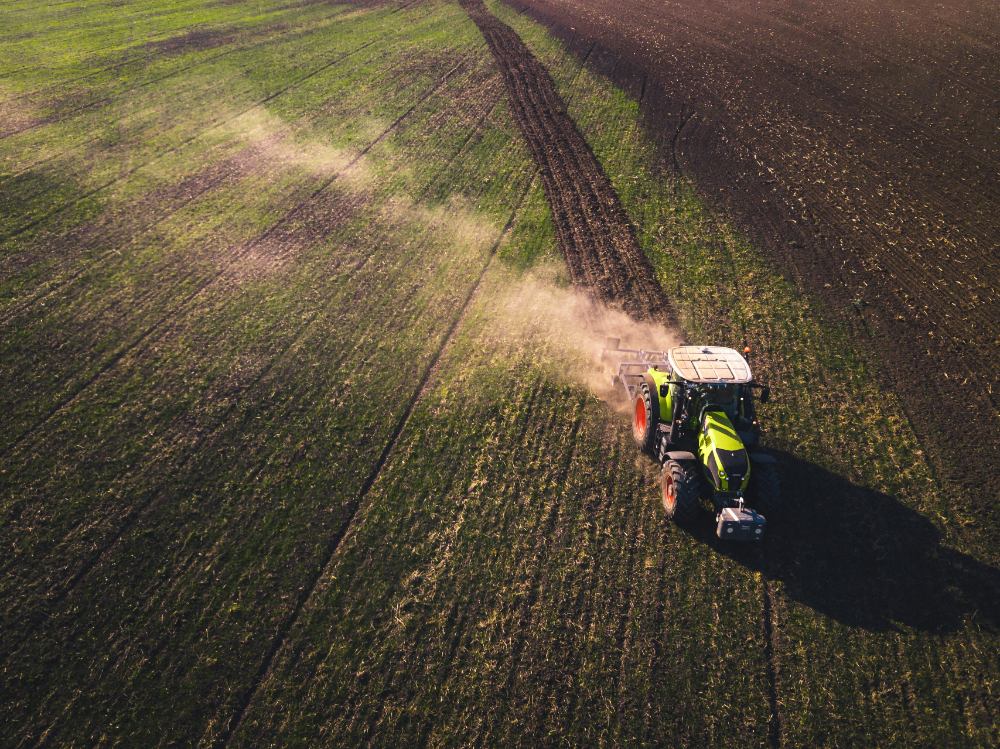 A tractor tilling the soil across a field, creating dust that catches the light from the sun