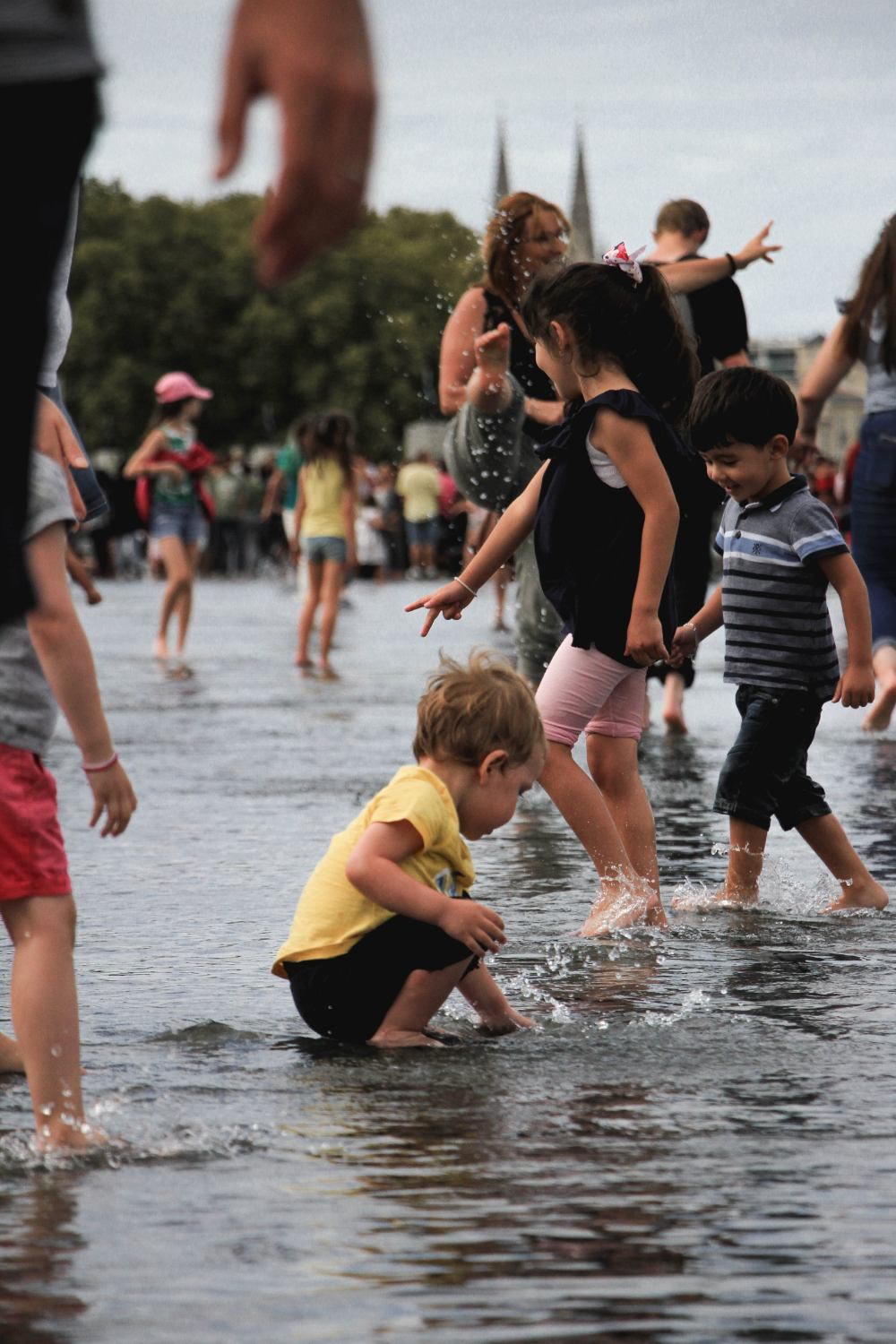 A toddler, crouched down and splashing his hand in shallow water while other children happily wet their feet and stroll around