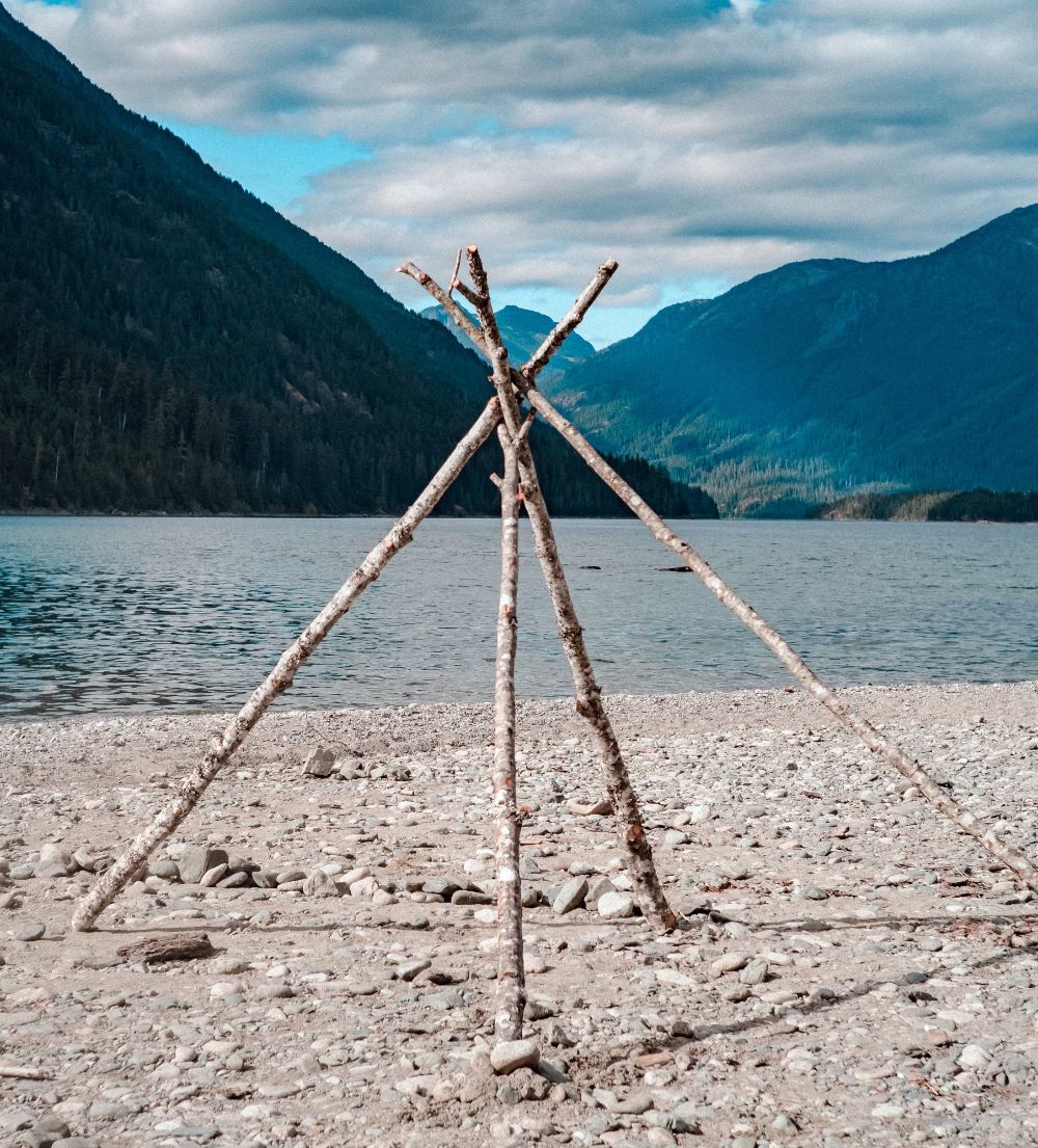 Four large sticks propped up in a pyramid shape on a rocky beach next to a lake and mountains